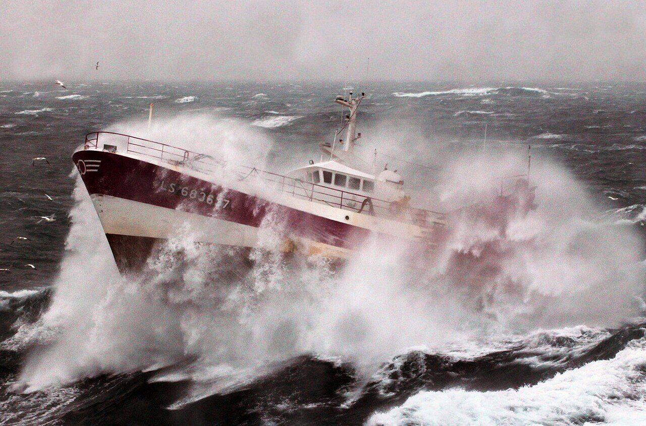 A small fishing vessel almost buried by a breaking wave in heavy seas.