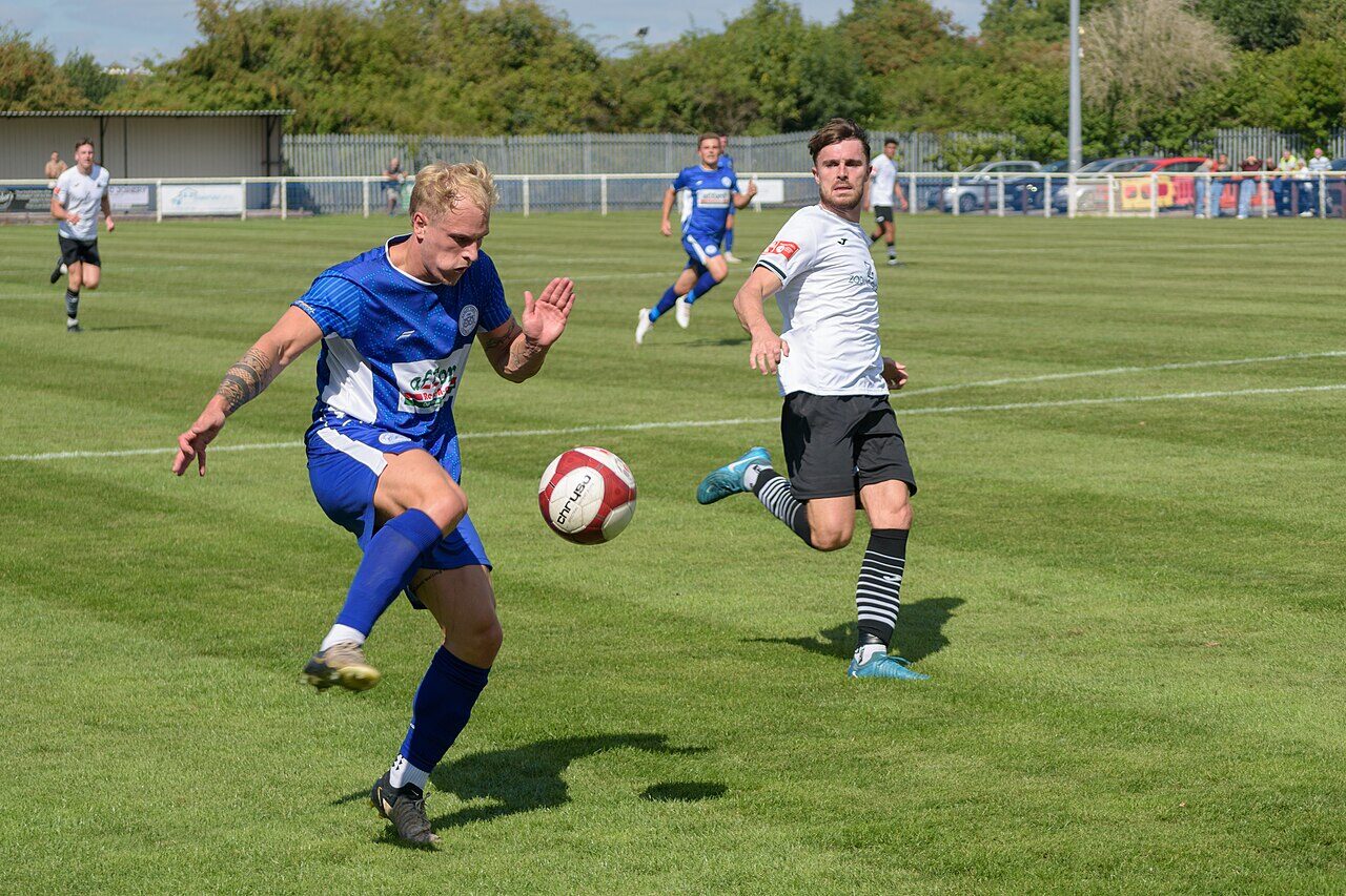 A footballer striking the ball during a match, with an opponent closing in.