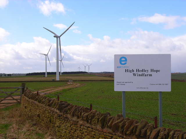 Wind turbines on open farmland behind a stone wall, with a sign reading High Hedley Hope Windfarm.