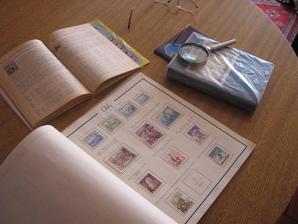 An open stamp album with a page of mounted Chilean stamps, beside a stamp catalogue and a magnifying glass.