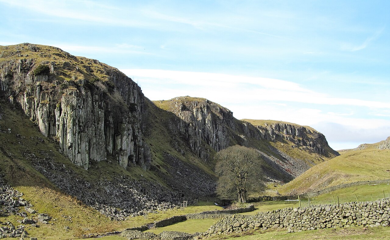 Grey cliffs of columnar dolerite rising above scree and drystone walls in a green valley.